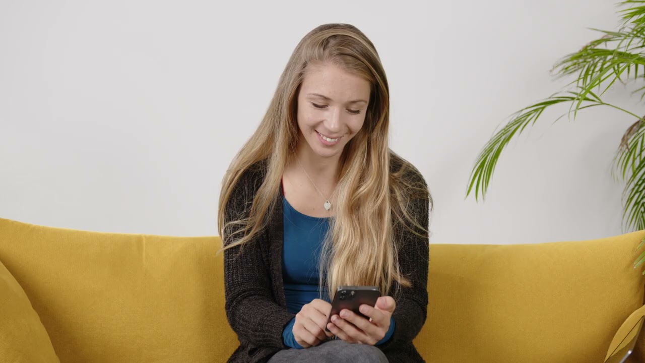 Frau mit langen blonden Haaren sitzend auf gelbem Sofa sieht auf Smartphone in ihren Händen